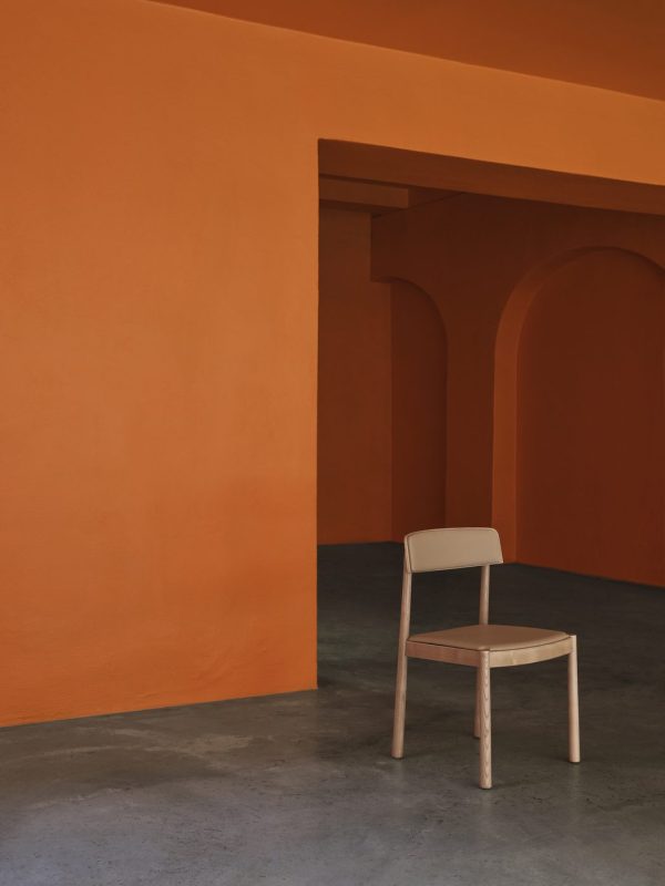 En enkelt beige designstol står op ad en levende orange væg, med en buet passage i venstre side, i et rum med blød belysning. Scenen understreger minimalisme og fed farvekontrast.