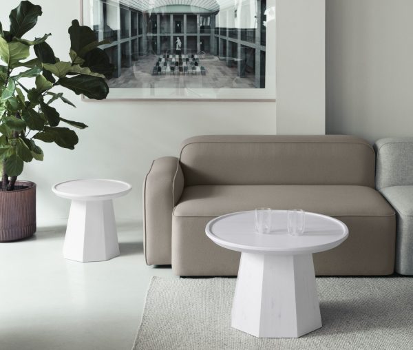 En moderne stue med en beige sofa, to hvide runde NORMANN COPENHAGEN - Pine Sofabord Ø65 borde, det ene med klare glas på, og et stort indrammet maleri af galleriudsigt. Der er også en stor grøn plante i hjørnet.