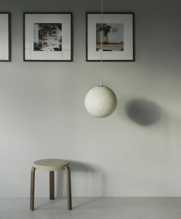 Et minimalistisk rum med et rundt, tekstureret NORMANN COPENHAGEN - Tap Skammel, der kaster en skygge på den grå væg, med tre indrammede billeder over en simpel skammel.