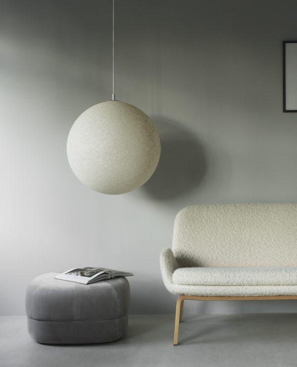 En minimalistisk stue med en tekstureret grå væg med en NORMANN COPENHAGEN - Pix Pendel Ø60 hængelampe, en moderne lysegrå sofa og en cirkulær grå skammel med en bog på. omgivelserne er rolige og stilfulde.