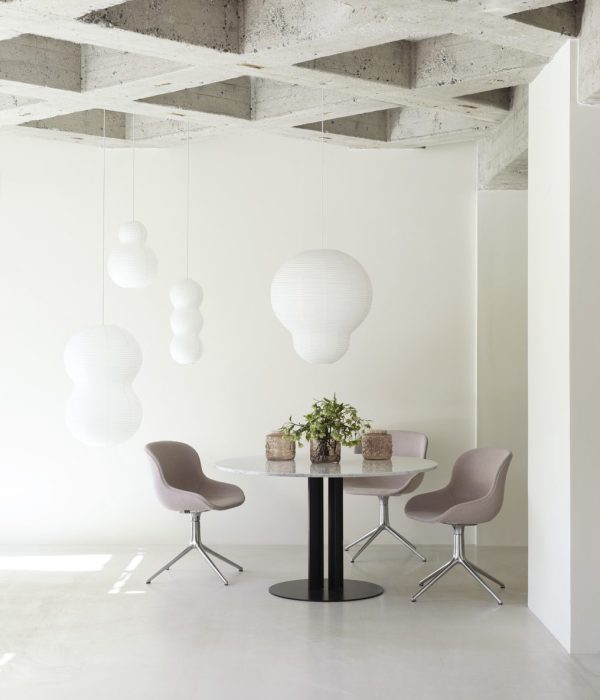 En minimalistisk spiseplads med et rundt bord og tre stole under unikke Normann Copenhagen pendellamper, på baggrund af synligt betonloft og hvide vægge.