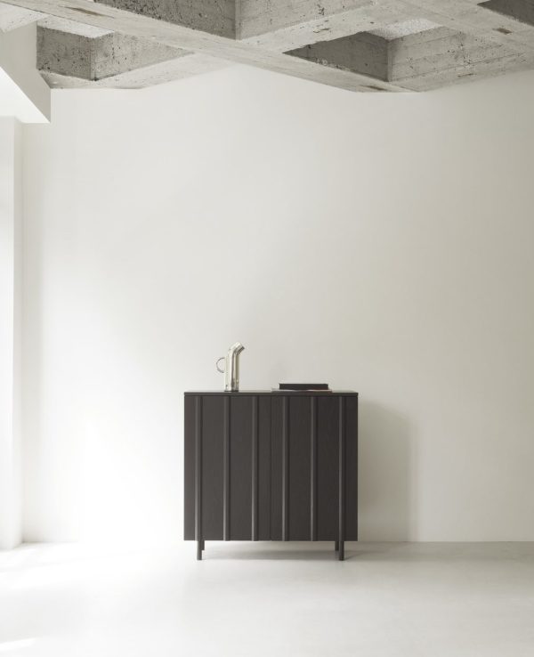 Et minimalistisk design med et sort skab med en vask og en vandhane sat mod en hvid væg og gulv. Rummet har et bart betonloft med synlige bjælker.