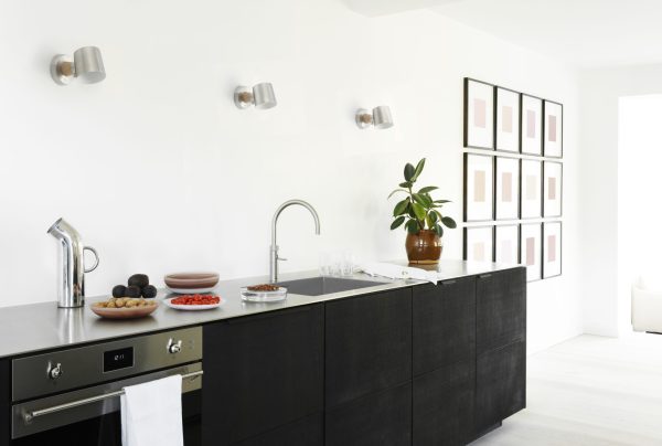 Moderne køkkenindretning med sorte skabe, hvidevarer i rustfrit stål og mørk bordplade. A NORMANN COPENHAGEN - Rise Væglampe hænger på en hvid væg, med planter og skåle med frugter på diskene.