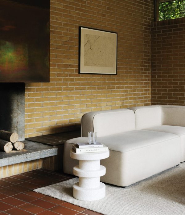 Et hyggehjørne i et værelse med en beige sofa og et unikt NORMANN COPENHAGEN - Bit Skammel Stack sidebord, placeret mod en synlig murstensvæg med pejs. Et indrammet abstrakt kunstværk hænger over pejsen.