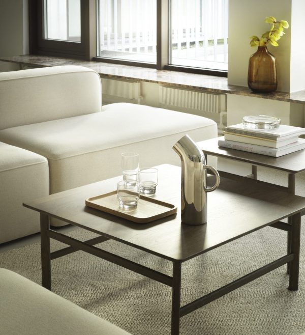 En hyggelig stue med en moderne beige sofa, et sofabord i træ med en bakke med glas og en NORMANN COPENHAGEN - Pipe Kande, bøger og en plante ved et vindue med persienner.