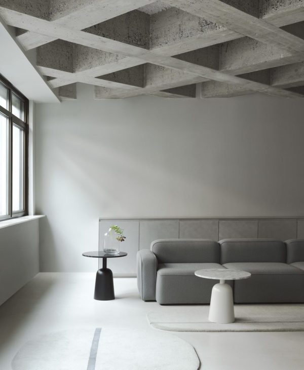 Moderne minimalistisk stue med en NORMANN COPENHAGEN - Rope Modulsofa 100 Narrow Venstre, et sort sidebord med en plante, en hvid vase på gulvet og synlige træbjælker i loftet. naturligt lys strømmer ind fra et vindue til venstre.