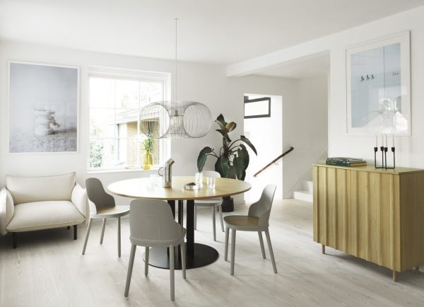En moderne spisestue med et rundt bord omgivet af fire stole, et træskab, store planter og vægmonteret kunst. Lyst rum med store vinduer og hvid indretning med TomorrowsDesign-elementer.