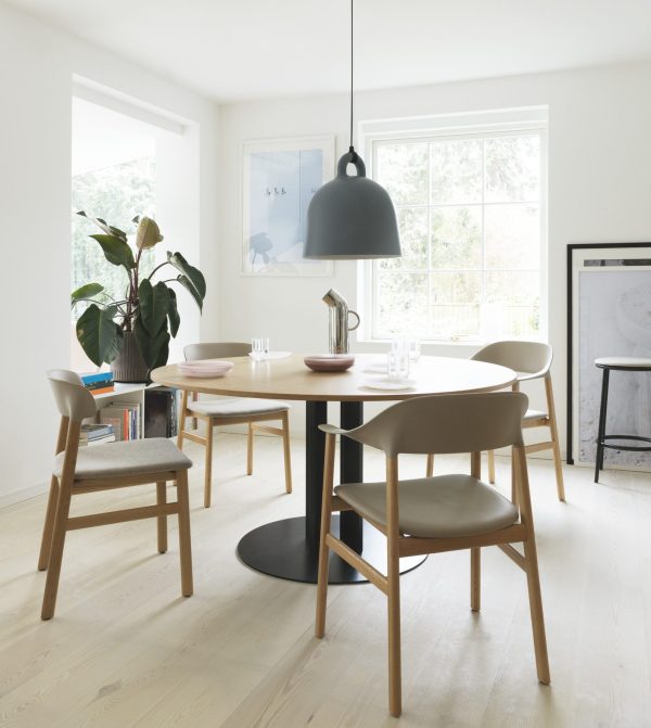 En minimalistisk spisestue med et rundt hvidt bord med møbler, en stor pendel og vinduer, der giver naturligt lys. En plante og indrammede kunstværker tilføjer dekorative detaljer.