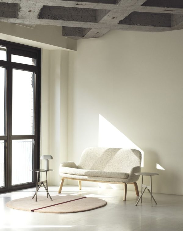 En minimalistisk stue med en cremet sofa, en NORMANN COPENHAGEN Stock Bord Ø37, et cirkulært tæppe og en sideskammel sat mod et stort vindue og tekstureret betonloft. stærkt sollys fylder rummet.
