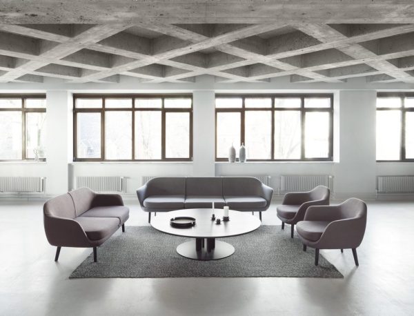 Moderne minimalistisk kontorlounge med TomorrowsDesign grå sofaer, to lyserøde lænestole, et hvidt rundt sofabord, sat på et gråt område med et synligt betonloft og store vinduer, der giver naturligt lys.