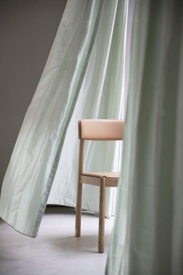 En minimalistisk træstol med et beige sæde foran et blødt grønt gardin, der er let åbent, sat i et rum med naturligt lys med TomorrowsDesign-interiør.
