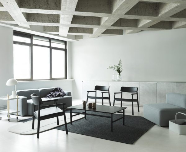 En minimalistisk stue med en grå sofa, to stole og et lille bord på et mørkt tæppe. Et sidebord, lampe og spredte hynder forbedrer rummet. Store vinduer giver naturligt lys, og synlige bjælker fremhæver loftet. Dette rum afspejler principperne for TomorrowsDesign med sin