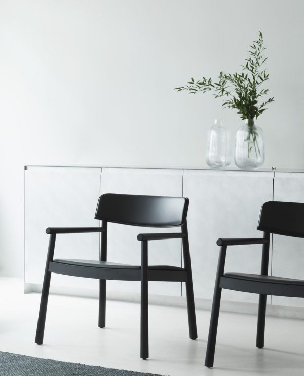 Et minimalistisk værelse med to sorte stole ud mod en hvid skænk. På skænken står tre klare vaser med grønne grene. Gulvet er gråt og væggen er hvid, hvilket understreger elegant designmøbler-æstetik.