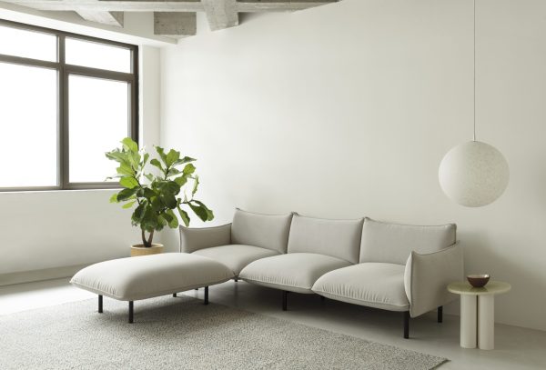 En minimalistisk stue med en NORMANN COPENHAGEN - Ark Modulsofa 100 Venstre, en rund, hvid hængelampe, en lille grøn plante på en beige skammel og en større potteplante nær et solbelyst vindue.