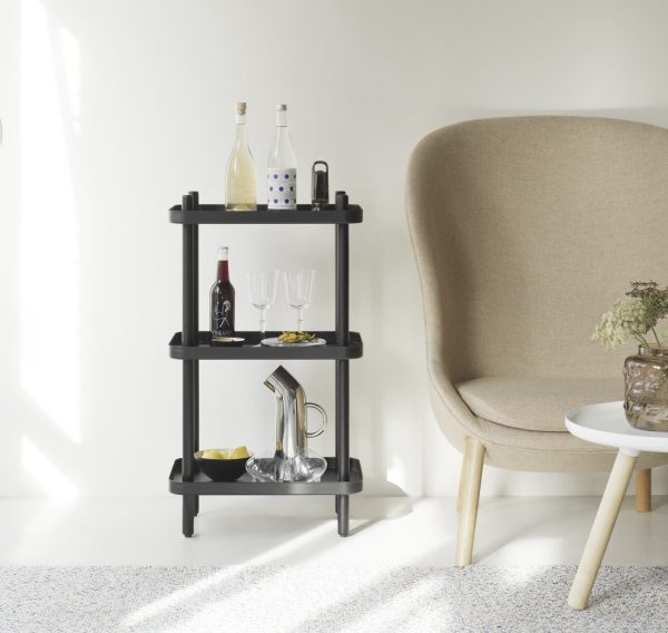 En moderne sort tre-lags NORMANN COPENHAGEN - Block Reol med forskellige flasker, glas og en dekorativ genstand i et lyst rum ved siden af en beige lænestol og et lille hvidt bord.