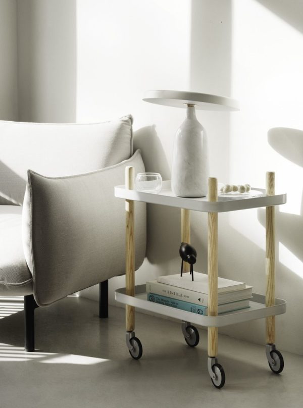 Et moderne stuehjørne badet i sollys, med en grå lænestol og et NORMANN COPENHAGEN - Block Rullebord med bøger, en lampe og pyntegenstande.
