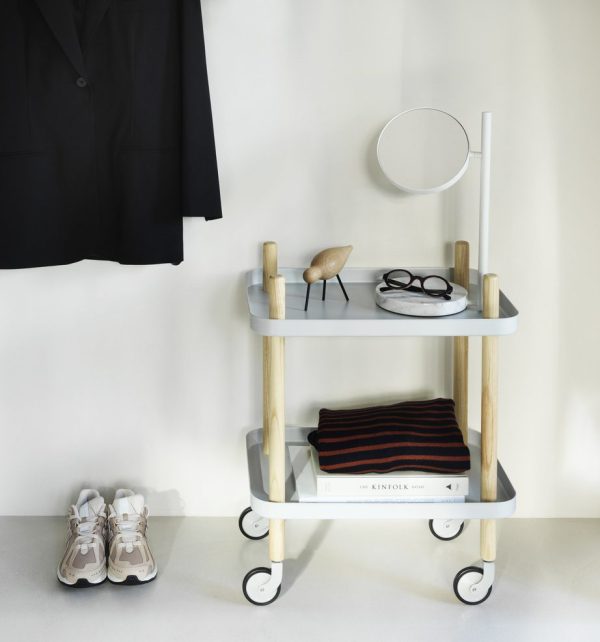 ET NORMANN COPENHAGEN - Rullebord med to hylder med forskellige ting: en sort jakke hængende på siden, briller, et lille rundt spejl, en fuglefigur i træ, bøger og sneakers placeret nedenunder.