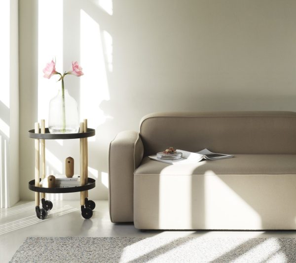 Et ryddeligt stuehjørne med sollys, der kaster skygger på væggen. En beige sofa, en NORMANN COPENHAGEN - Block Rullebord Ø45 med en vase med lyserøde blomster og et par bøger skaber en rolig atmosfære.