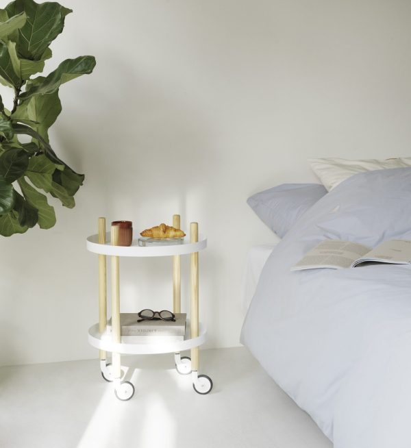 En lille, hvid design serveringsvogn med morgenmadsartikler ved siden af en uopredt seng med en hvid dyne. Vognen rummer en kaffekop, croissant, bog og briller i et neutralt tonet rum med sollys.