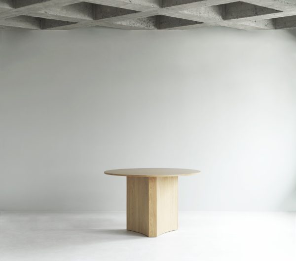 Et minimalistisk rum med et groft, synligt betonloft og en almindelig grå væg, med et enkelt rundt træbord centreret på et lysegråt gulv, afspejler slanke designprincipper.