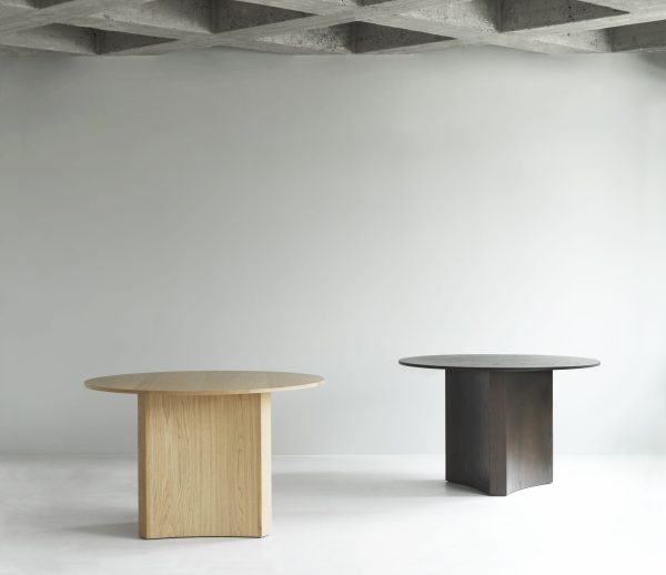 To minimalistiske designmøbler-borde i et almindeligt rum, et lyst træ og et mørkt, placeret på et betongulv mod en hvid væg med synlige overliggende bjælker.