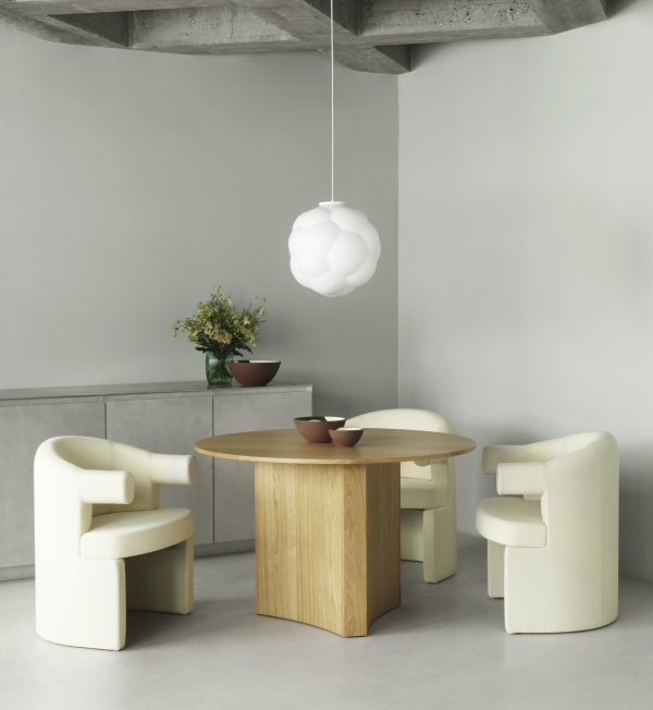 En moderne spiseplads med et rundt træbord og fire cremefarvede lænestole, med designmøbler, under en hvid, skyformet pendel, i et rum med betonvægge og gråt gulv.