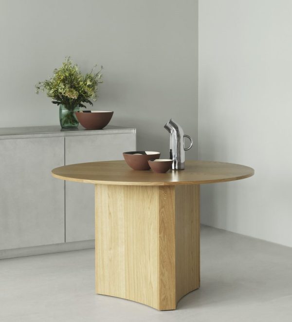 Et minimalistisk køkken med et rundt træbord med indbygget NORMANN COPENHAGEN - Pipe Kande og vandhane. Bordet rummer en skål, en tallerken og en vase med blomster. Baggrunden viser en almindelig grå væg og en grå tæller.