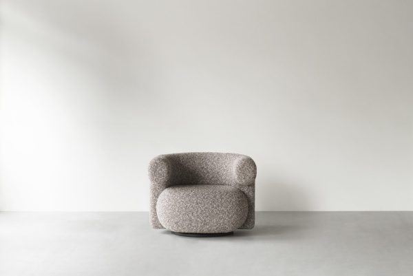 Et moderne, gråt, tekstureret stof NORMANN COPENHAGEN - Burra Loungestol M. Drejefunktion lænestol placeret i midten af et minimalistisk rum med grå vægge og gulve, oplyst af naturligt lys, der skaber en blød skygge.