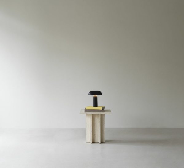 En minimalistisk komposition med en NORMANN COPENHAGEN - Porta Bordlampe i et neutralt tonet rum med en sort cirkulær genstand på en gul bog. baggrunden er en almindelig lysegrå væg.