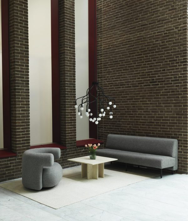 En moderne stue med en grå sofa, en cylindrisk grå stol og et sofabord i træ med en vase med blomster. Rummet har murstensvægge, lodrette hvide og bordeaux striber og et stilfuldt design hængende lysarmatur.