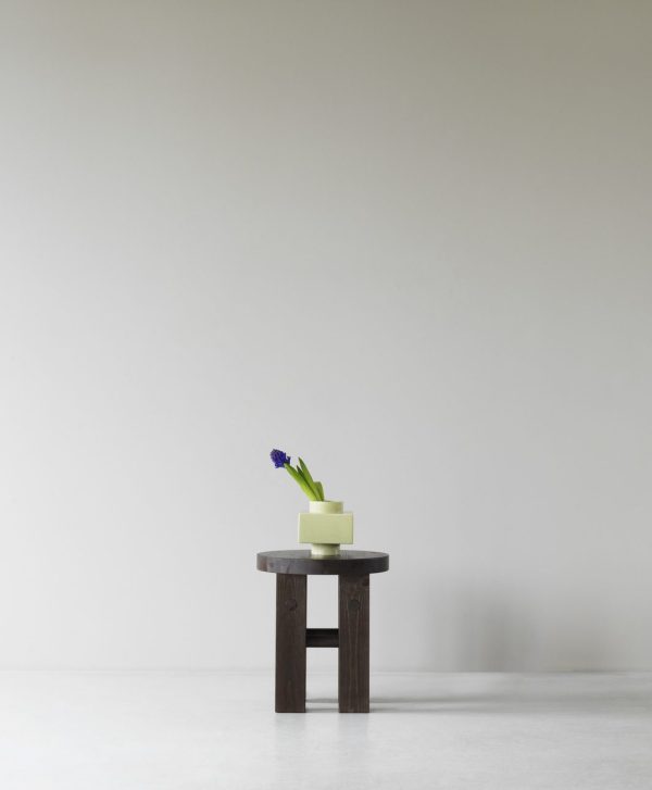 Et minimalistisk setup med NORMANN COPENHAGEN - Fyr Skammel i et rum med neutral baggrund. På toppen af skamlen er en firkantet, lysegrøn vase med en enkelt blå blomst.