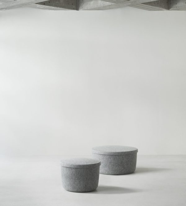 To runde, grå NORMANN COPENHAGEN - Skjul Opbevaringspuf Stor taburetter i et tomt rum med hvide vægge og betongulv. loftet har synlige træbjælker. scenen kommunikerer enkelhed og minimalisme.