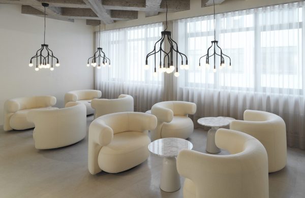 Moderne minimalistisk loungeområde med hvide skulpturelle lænestole og runde sideborde i marmor, oplyst af unikke loftslamper. Naturligt lys filtrerer gennem rene gardiner i et rummeligt rum med betonloft. Interiøret afspejler en sofistikeret blanding af form og funktion.