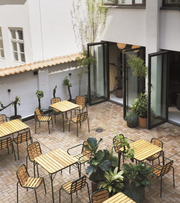 En rolig gårdhavecafé med NORMANN COPENHAGEN - Vig Bord 90X80 borde og stole omgivet af frodige potteplanter. store, åbne døre med sort ramme forbinder rummet med et indendørs område, hvilket forstærker den hyggelige, indbydende stemning.
