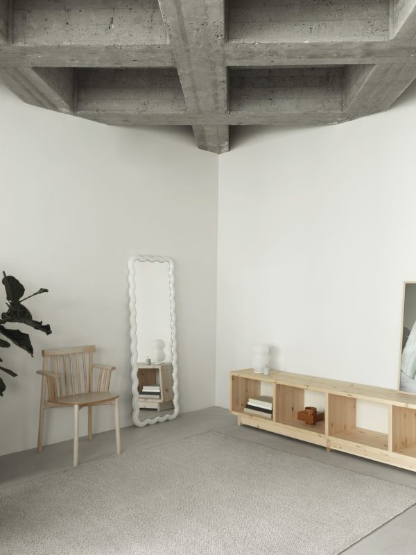 En minimalistisk stue med en træstol ved siden af et unikt bølget spejl, en lav træhylde med bøger og opbevaringskasser, et stort tæppe og en potteplante ved siden af synlige betonbjælker viser TomorrowsDesign.