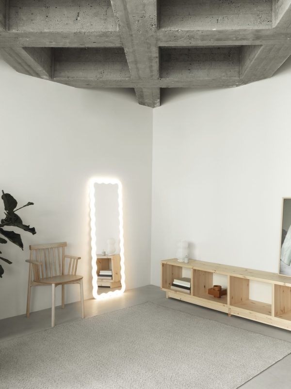 Et moderne minimalistisk opholdsrum med en NORMANN COPENHAGEN - Plank Bogreol Lav med indbygget belysning, en træstol, et lavt opbevaringsrum og en plante. rummet har synlige betonlofter og et tæppe på gulvet.
