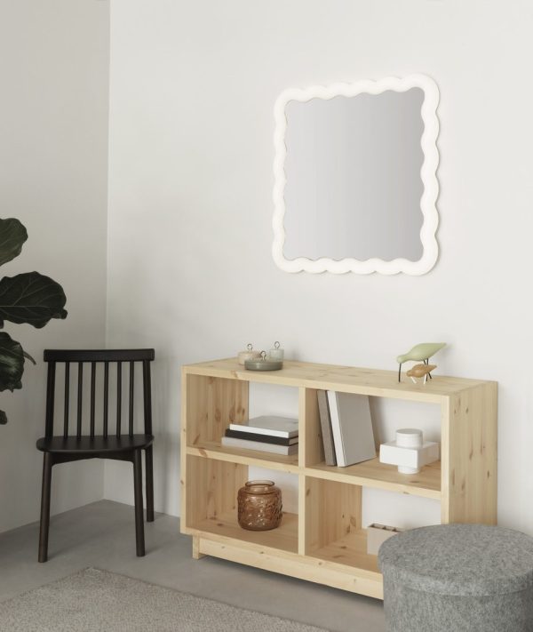 Et minimalistisk værelse med en NORMANN COPENHAGEN - Plank Bogreol Mellem indeholdende bøger og pynteting, en mørk stol, en grå puf og et karakteristisk bølgekantet spejl på væggen.