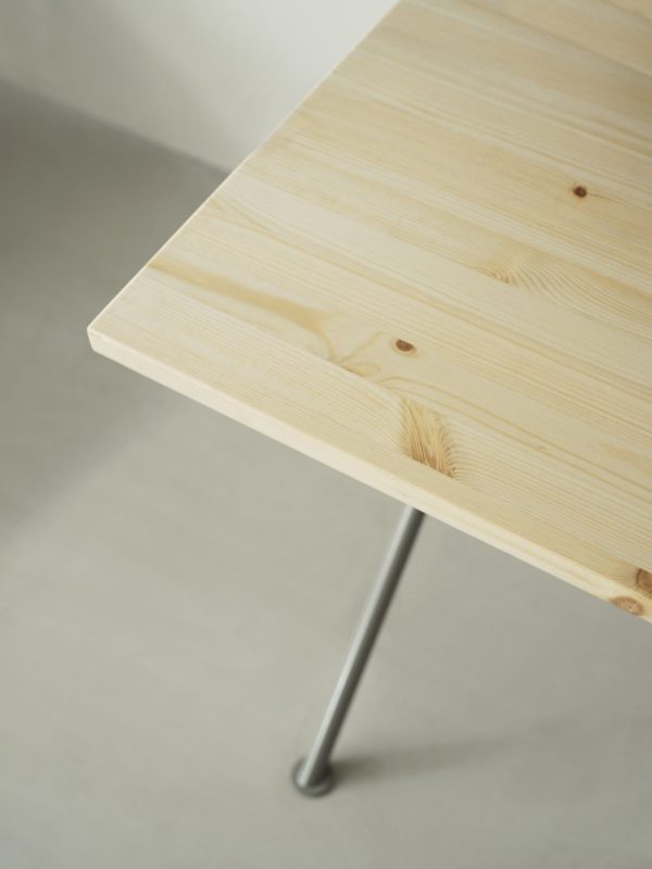 Et nærbillede af et NORMANN COPENHAGEN - Kip Skrivebord med en glat, let træoverflade med synlige årer og knaster, understøttet af et enkelt metalben. baggrunden er blødt oplyst og neutral farvet.
