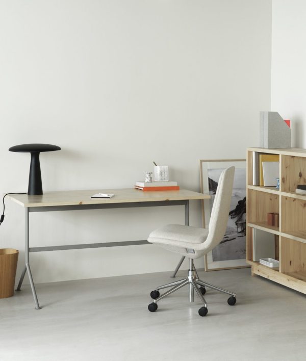 Et minimalistisk arbejdsområde med et NORMANN COPENHAGEN - Kip Skrivebord skrivebord med en sort lampe og en notesbog, en beige kontorstol og en reol i træ med bøger og en skraldespand mod en almindelig, grå væg med hvidt gulv.