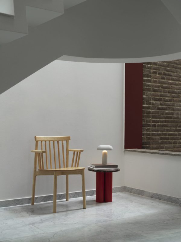 Et minimalistisk interiør med en enkelt træstol og et lille rødt bord med en moderne, stearinlyslignende lampe på. Indstillingen omfatter en hvid væg, en murstensvæg synlig gennem et buet vindue og et marmorgulv. Dette designmøbler setup eksemplificerer moderne elegance blandet med traditionel arkitektur