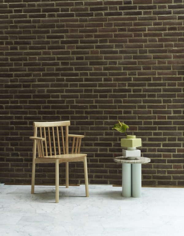 En træstol og en stak NORMANN COPENHAGEN - Deko Object S4 ved siden af, placeret mod en mørk murstensvæg med marmorgulv. en lille plante sidder på toppen af cylindrene.
