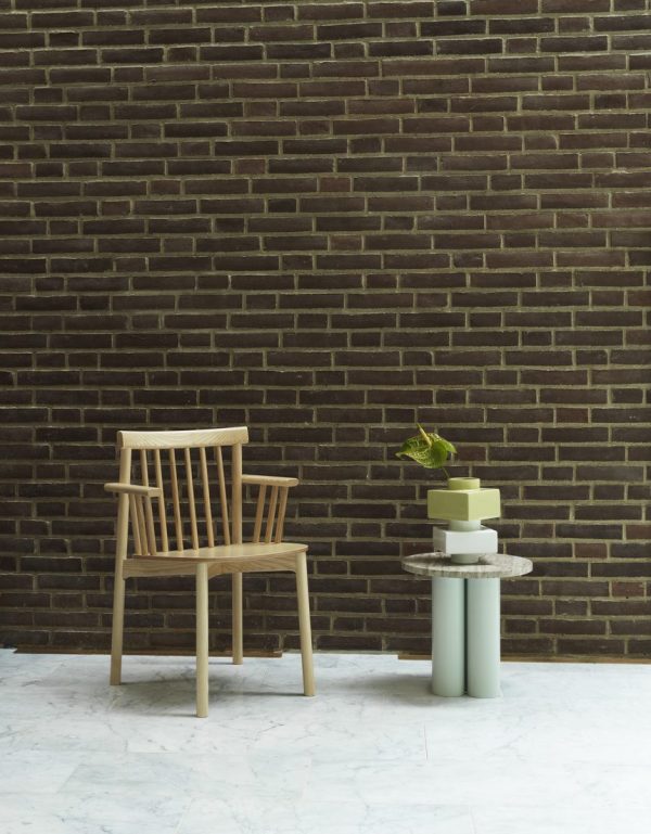 En træstol ved siden af en stak farverige cylindriske genstande på et lille bord, placeret op ad en mørk murstensvæg, alt sammen sat på et marmorgulv med designmøbler.