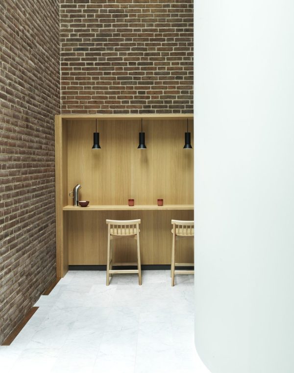 Et minimalistisk caféhjørne med træstole og træbord, indbyggede hylder og moderne pendellamper. Røde krus fremhæver murstensvæggenes neutrale toner og lette designmøbler.