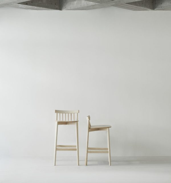 To træstole, den ene en barstol og den anden en standard spisestuestol, står side om side mod en almindelig hvid væg i minimalistiske omgivelser med designelementer og et synligt betonloft.