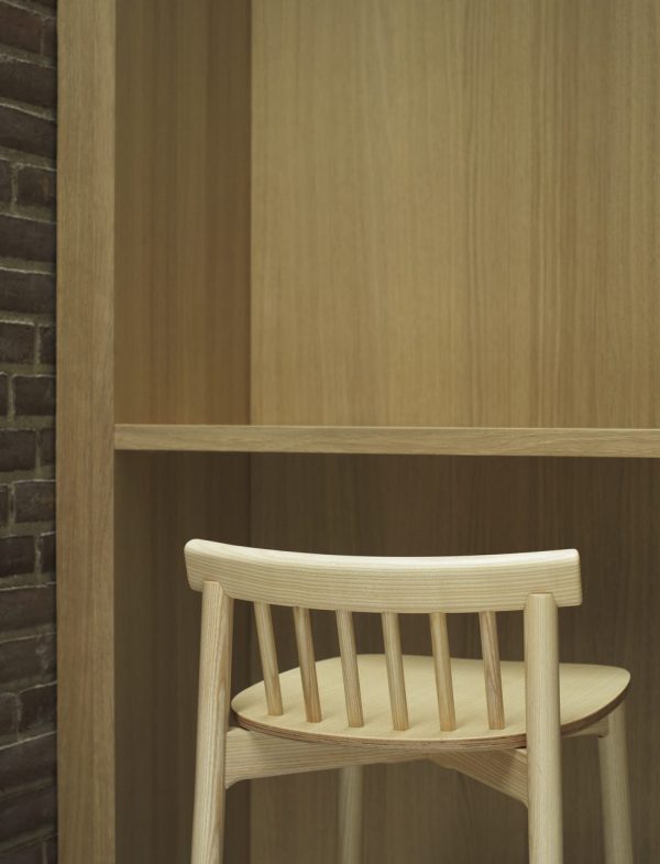 En træstol med buet ryglæn placeret mod en matchende træbogreol i minimalistiske omgivelser. De anvendte materialer udviser en naturlig, let træfinish, der afspejler en skandinavisk designmøbler-æstetik.