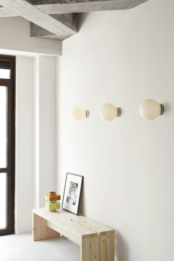 Tre NORMANN COPENHAGEN - Pix Lampe Ø20 er monteret på en hvid væg over en træbænk med dekorative genstande og et indrammet billede, nær et vindue i et rum med synlige betonbjælker.