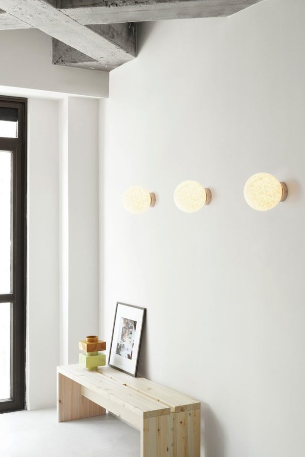 Et minimalistisk interiør med en hvid væg med tre runde, teksturerede NORMANN COPENHAGEN - Stretch Bænk 160 lysarmaturer. Under lysene er en træbænk med et par dekorative genstande og et indrammet billede ved siden af et vindue.
