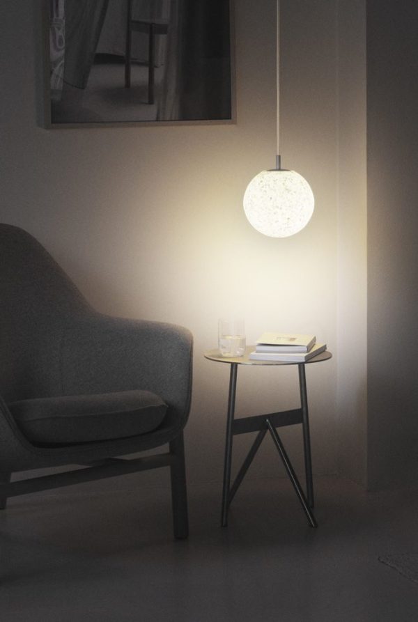 Et hyggeligt hjørne af et værelse med en grå lænestol ved siden af et lille, trekantet sidebord med en bog og et glas på. Over bordet udsender en NORMANN COPENHAGEN - Pix Pendel Ø20 en blød glød.