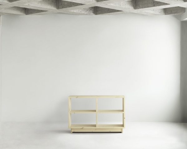 Et minimalistisk interiør, der viser en NORMANN COPENHAGEN - Plank Bogreol Mellem i et sparsomt møbleret rum med gråt betonloft og gulv, oplyst af blødt diffust lys fra højre side.
