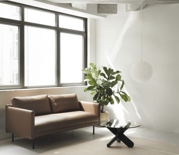 En moderne stue med en solbrun lædersofa, et rundt glas sofabord med sort bund, en stor potteplante og en NORMANN COPENHAGEN - Pix Pendel Ø30. rummet har store vinduer og hvide vægge.